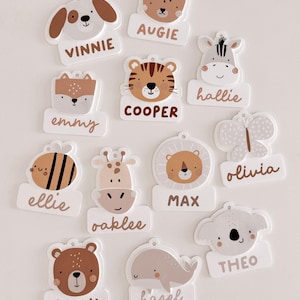 May include: A selection of animal name tags. Each tag features a different animal illustration, such as a dog, bear, tiger, zebra, fox, bee, giraffe, lion, butterfly, koala, and whale. Each tag has a name written below the animal.