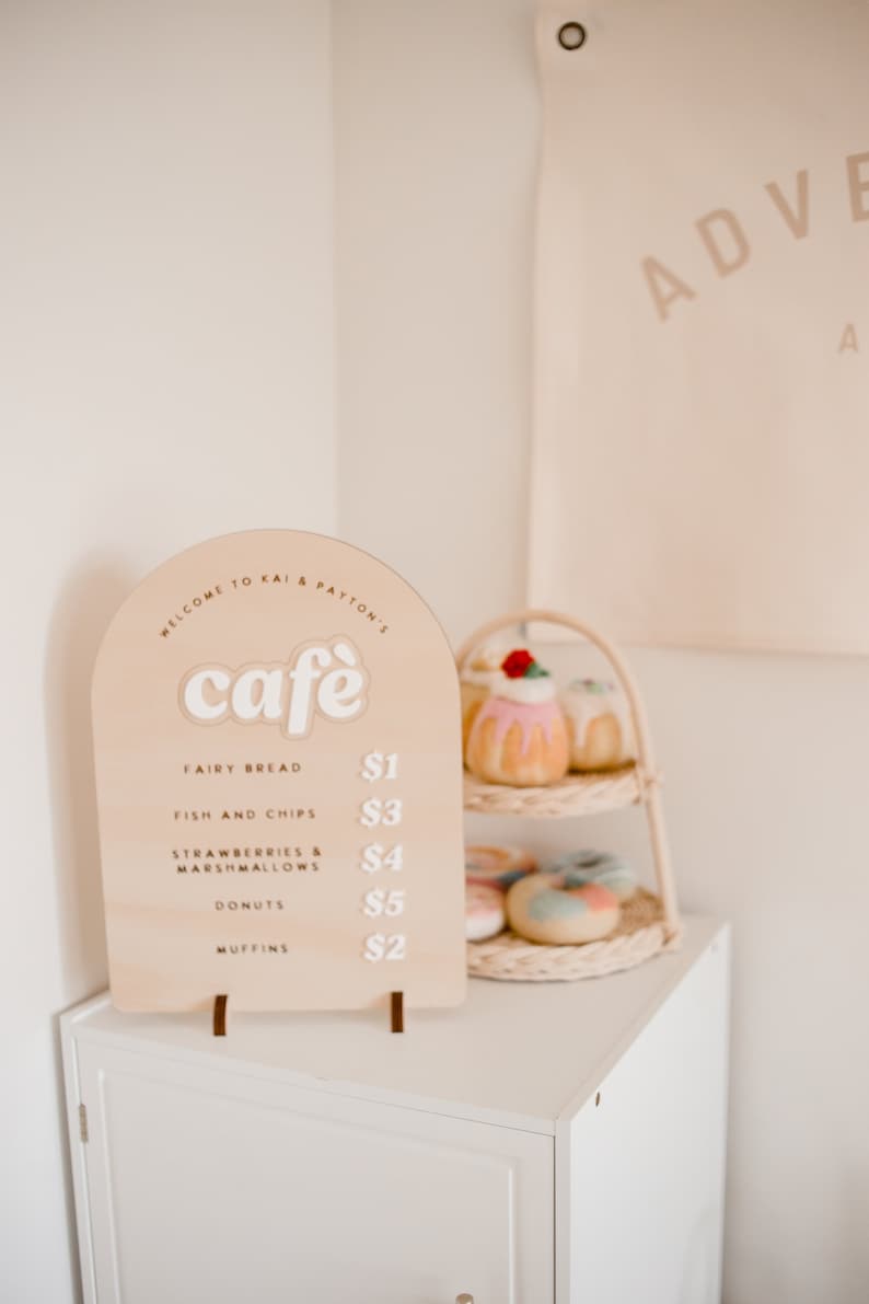Pu&ograve; includere: Un cartello in legno con un menu di caffetteria per un caff&egrave; di gioco per bambini. Il cartello ha la forma di un arco e in cima ha le parole "Benvenuti al caff&egrave; di Kai & Payton". Il menu elenca articoli come pane fatato, pesce e patatine fritte, fragole e marshmallow, ciambelle e muffin con prezzi accanto a ogni articolo.