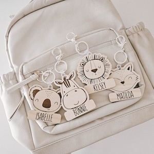 Può includere: Cinque portachiavi in legno con volti di animali e nomi personalizzati. I portachiavi presentano un leone, una zebra, un koala, una volpe e un'etichetta con i nomi Kelsey, Bonnie, Isabelle, Matilda.