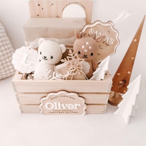 May include: A wooden crate filled with Christmas-themed toys and decorations. Includes a teddy bear and a deer plush, a wooden Christmas tree, and a sign that says "Oliver". The crate is decorated with a snowflake and a tag that says "Santa's Bag".