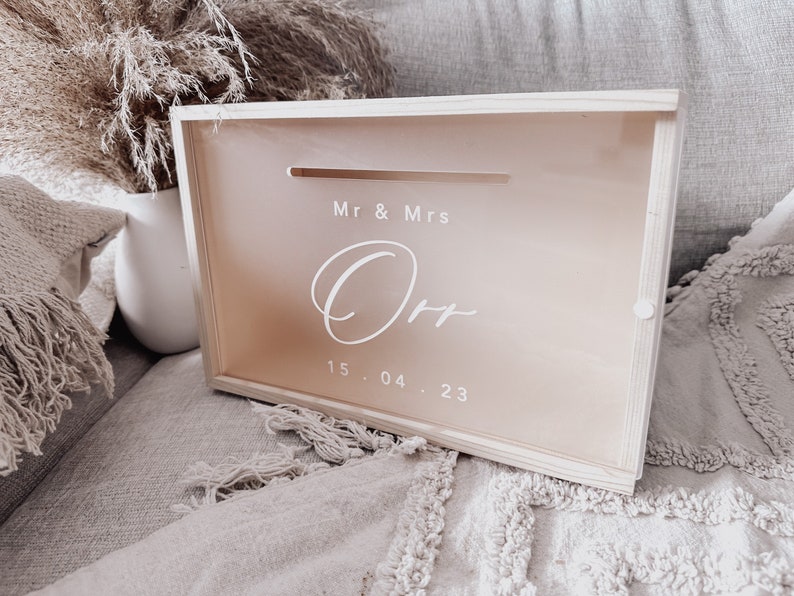 Puede incluir: Una caja de madera con una ranura en la parte superior para tarjetas o dinero. La caja est&aacute; decorada con el texto "Mr & Mrs Orr" y la fecha "15.04.23".