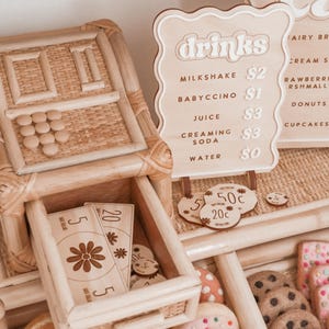 May include: A wooden play food stand with a drinks menu, play money, and various felt food items. The menu lists drinks like milkshake and juice. The stand includes a cash register and a drawer with play money.