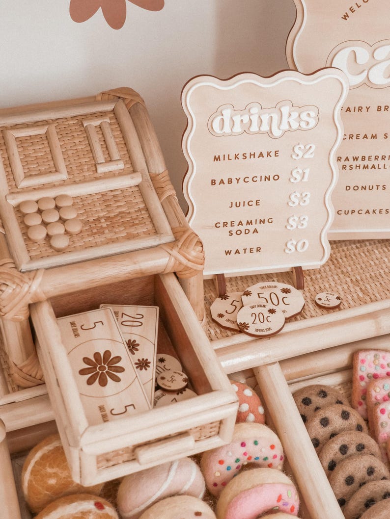 May include: A wooden play food stand with a drinks menu, play money, and various felt food items. The menu lists drinks like milkshake and juice. The stand includes a cash register and a drawer with play money.