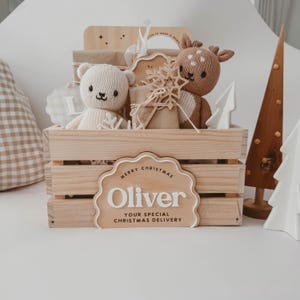 May include: A wooden crate filled with Christmas gifts, a teddy bear, and a deer plushie. The crate has a sign that reads "Merry Christmas Oliver, Your Special Christmas Delivery". A wooden Christmas tree and a white tree are on the right.