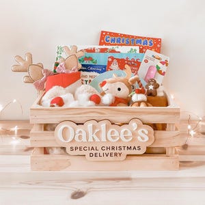 Può includere: Una cassa di legno piena di giocattoli e libri a tema natalizio. La cassa ha la scritta "Oaklee's Special Christmas Delivery" sul davanti. All'interno ci sono renne di peluche, un omino di pan di zenzero e vari libri.