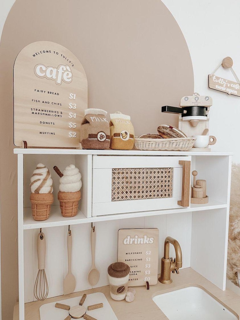 Pu&ograve; includere: Un set da cucina in legno a tema caff&egrave;. La cucina presenta un armadio bianco con un cestino in vimini marrone, un lavandino con un rubinetto dorato e un piano cottura. Ci sono due cartelli in legno con il testo "Benvenuti al Tully's cafe" e "Tully's cafe ora aperto". La cucina include anche una macchina da caff&egrave; in legno, due cartoni di latte e due coni gelato.