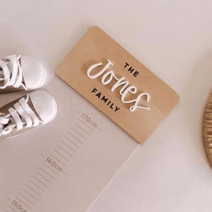 Può includere: Una targa in legno chiaro con il testo THE JONES FAMILY in lettere bianche e nere, posizionata accanto a un paio di scarpe da neonato beige con lacci bianchi e un metro a parete con misurazioni fino a 170cm.