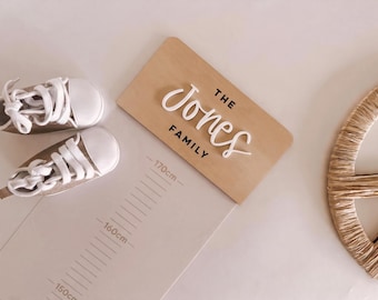 May include: A light-colored wooden plaque with the text THE JONES FAMILY in white and black lettering, positioned next to a pair of beige baby sneakers with white laces and a height chart marked with measurements up to 170cm.
