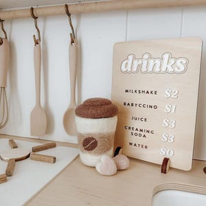 Pu&ograve; includere: Un cartello di legno con la parola "bevande" in lettere bianche su sfondo marrone. Il cartello elenca diverse bevande e i loro prezzi: milkshake $2, babyccino $1, succo $3, soda cremosa $3, acqua $0. Una tazza di caff&egrave; in feltro con un coperchio marrone e un design a chicco di caff&egrave; marrone si trova su un bancone di legno.
