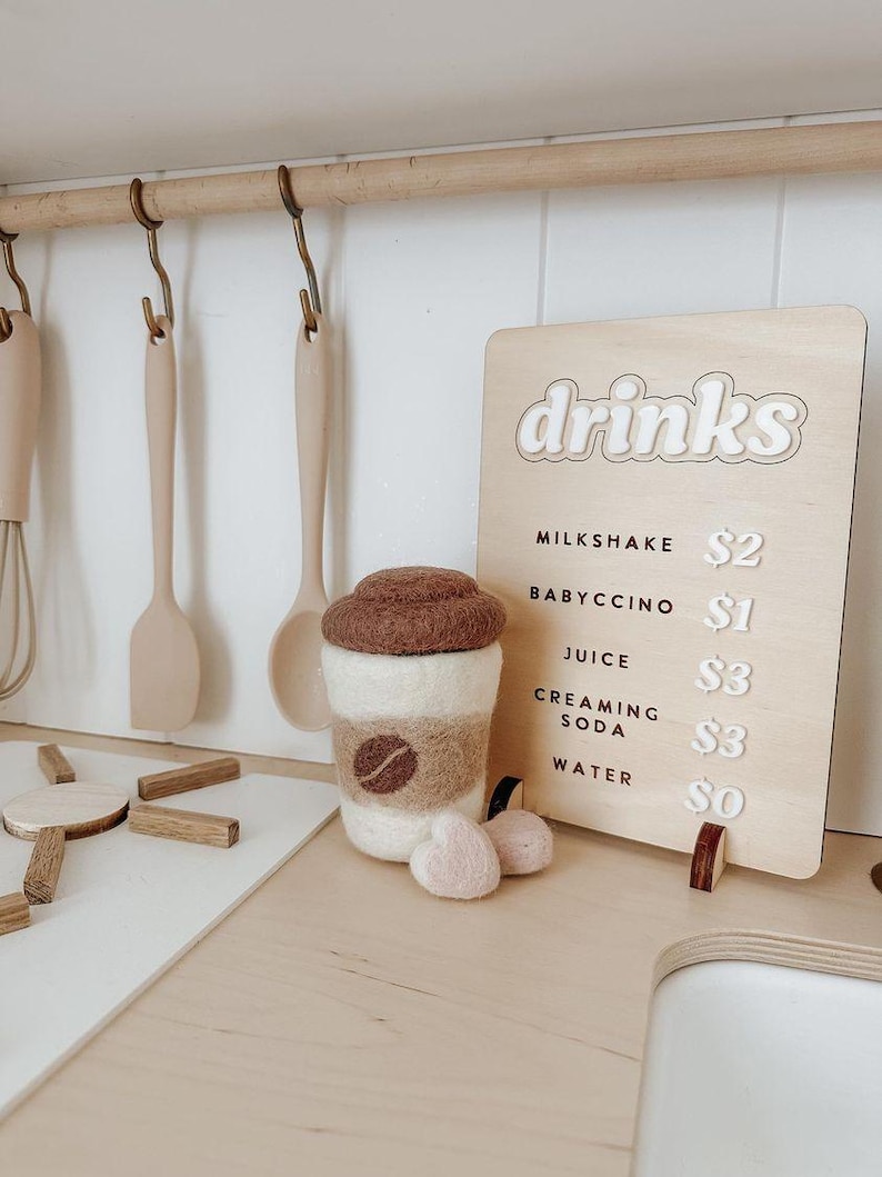 Pu&ograve; includere: Un cartello di legno con la parola "bevande" in lettere bianche su sfondo marrone. Il cartello elenca diverse bevande e i loro prezzi: milkshake $2, babyccino $1, succo $3, soda cremosa $3, acqua $0. Una tazza di caff&egrave; in feltro con un coperchio marrone e un design a chicco di caff&egrave; marrone si trova su un bancone di legno.