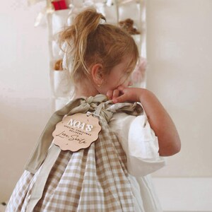 May include: A tan and white checkered gift bag with a wooden tag that reads "NOA'S SPECIAL CHRISTMAS DELIVERY Love, Santa". The bag is tied with a tan ribbon. The bag is being held by a person wearing a white dress.