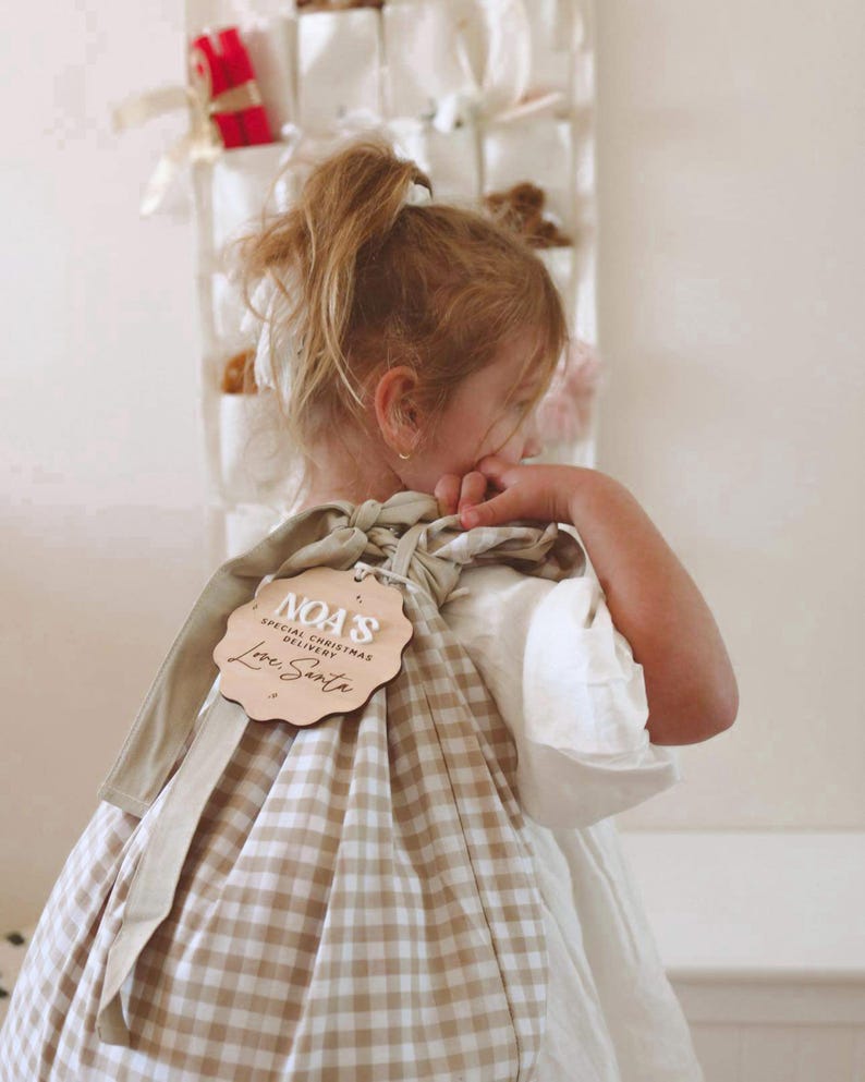 May include: A tan and white checkered gift bag with a wooden tag that reads "NOA'S SPECIAL CHRISTMAS DELIVERY Love, Santa". The bag is tied with a tan ribbon. The bag is being held by a person wearing a white dress.