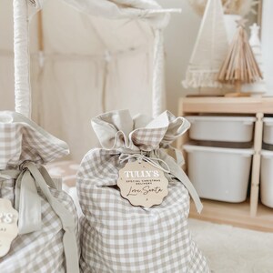 May include: Two beige and white gingham fabric gift bags with drawstring closures. Each bag has a wooden tag that reads "Tully's Special Christmas Delivery Love, Santa". The bags are tied with a beige ribbon.