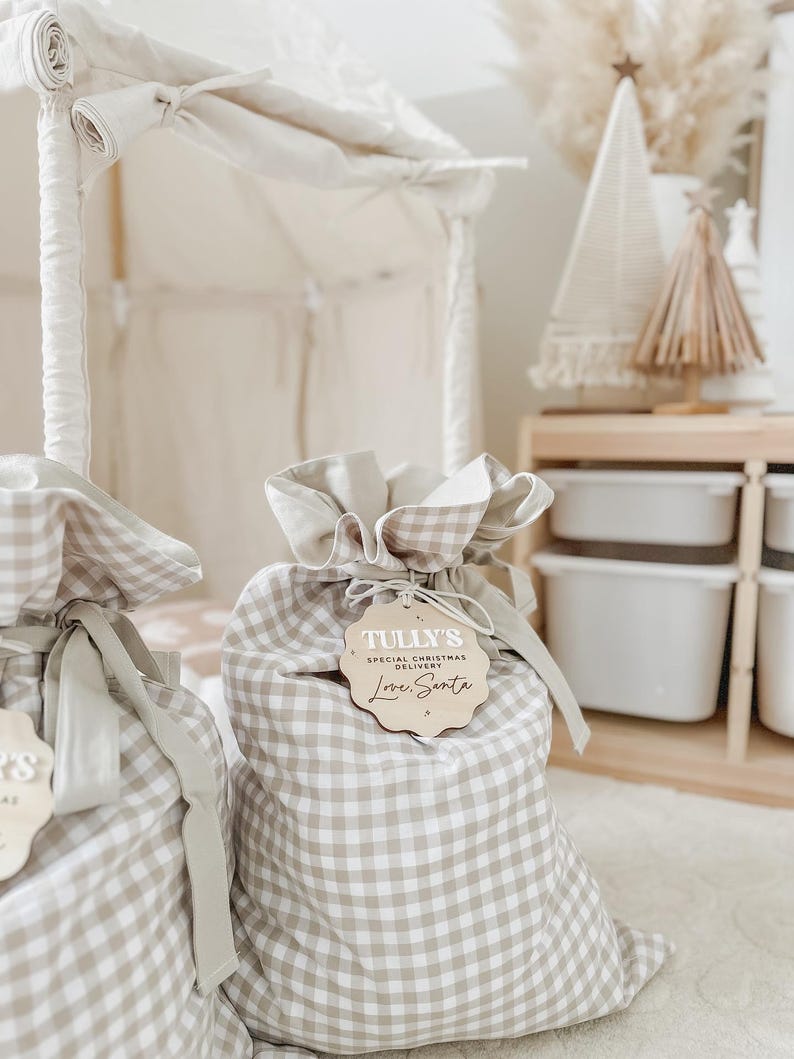 May include: Two beige and white gingham fabric gift bags with drawstring closures. Each bag has a wooden tag that reads "Tully's Special Christmas Delivery Love, Santa". The bags are tied with a beige ribbon.