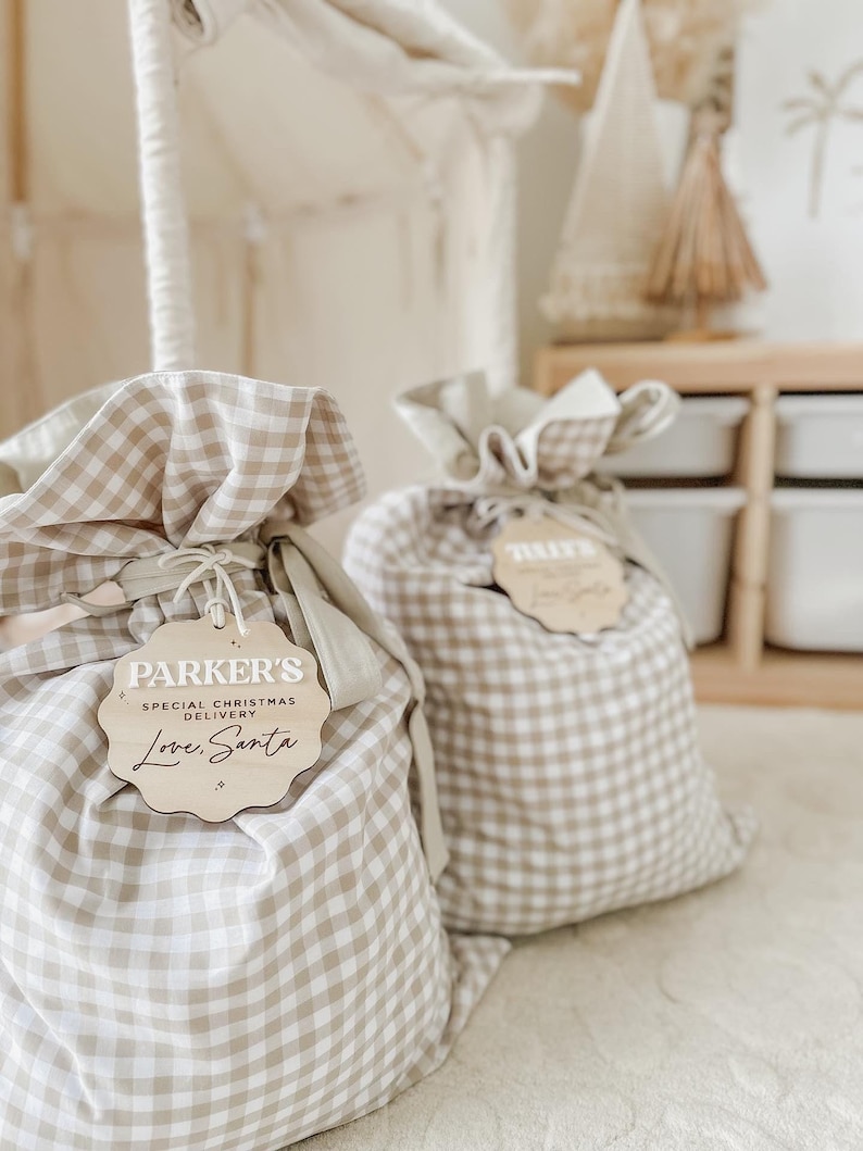 May include: Two beige and white gingham fabric gift bags with wooden tags. The tags read "Parker's Special Christmas Delivery Love, Santa". The bags are tied with a natural twine and are sitting on a cream-colored surface.