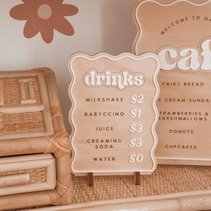 May include: Wooden cafe menu sign with the word "drinks" in white, listing milkshake, babyccino, juice, creaming soda, and water. The sign is on a wooden stand, with a flower design on the wall.