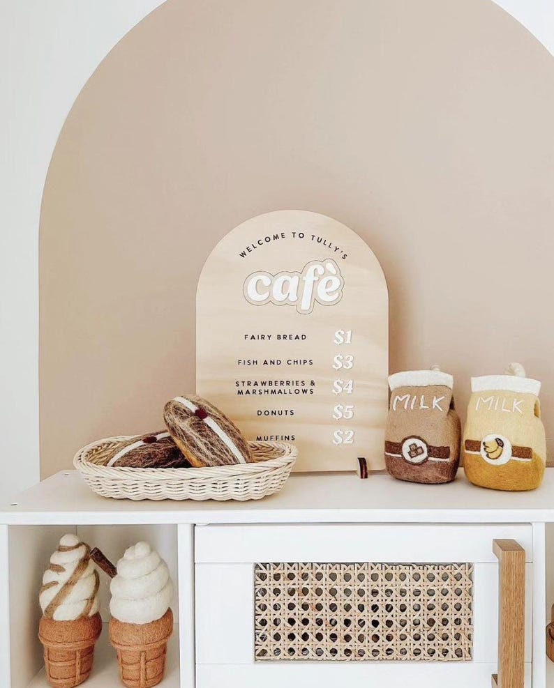 Pu&ograve; includere: Un cartello di legno con le parole "Benvenuti al Tully's cafe" e un menu che elenca i prezzi per il pane delle fate, il pesce e le patatine fritte, le fragole e i marshmallow, i donut e i muffin. Il cartello &egrave; su un bancone bianco con un cestino di pasticcini di feltro marrone e bianco e due cartoni di latte di feltro.