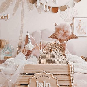 May include: A wooden crate filled with decorative items, including a unicorn horn headband, a star-shaped headpiece, and a woven purse. A sign reading "Merry Christmas Isla" is on the front. The scene is set in a room with a palm tree mural.