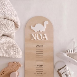 May include: A light-coloured wooden growth chart with an arched top, featuring a white 3D dinosaur silhouette and the name NOA in white lettering. The chart displays measurement markings from 180cm to 200cm in dark brown ink on the smooth wood surface.