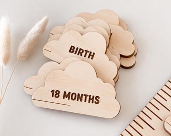 May include: A stack of light-colored wooden cloud-shaped plaques featuring dark engraved text including BIRTH and 18 MONTHS. Each plaque has a horizontal line for writing. The set is arranged with two dried grass stems and a wooden ruler edge visible.