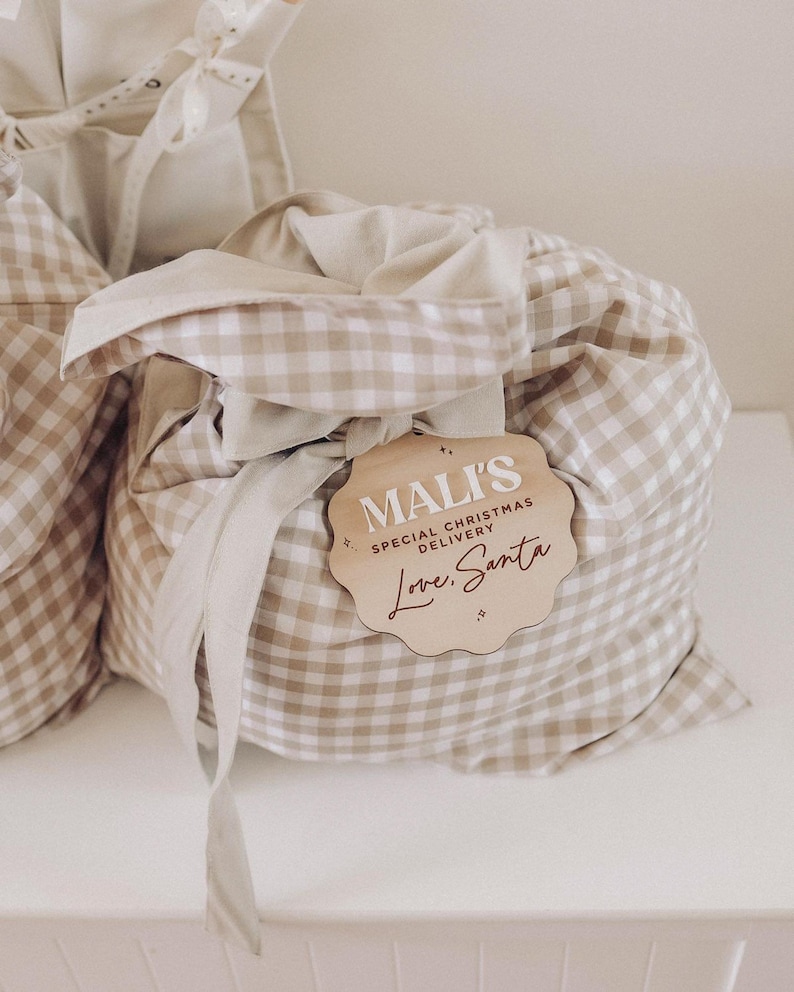 May include: Tan and white gingham fabric gift bag with a matching ribbon and a wooden tag. The tag reads "Mali's Special Christmas Delivery Love, Santa" in a decorative font. The bag is tied closed.