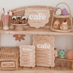 May include: A charming display of felt food and cafe signage. The sign reads "Oaklee's Cafe, Est. 2024". Menu boards list drinks and treats like milkshakes, cupcakes, and donuts. The decor includes a woven shelf and flower accents.