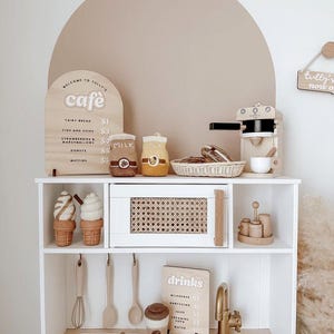 Pu&ograve; includere: Una cucina giocattolo bianca con un piano di lavoro in legno marrone e un lavandino. La cucina ha un piccolo forno, una macchina per il caff&egrave; e uno scaffale con un cartello che dice "Benvenuti al Tully's Cafe". Ci sono anche due coni gelato di feltro sullo scaffale.