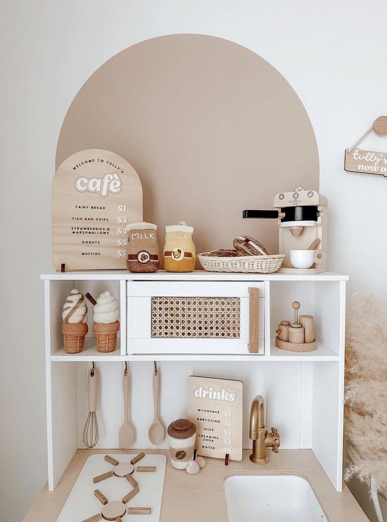 Pu&ograve; includere: Una cucina giocattolo bianca con un piano di lavoro in legno marrone e un lavandino. La cucina ha un piccolo forno, una macchina per il caff&egrave; e uno scaffale con un cartello che dice "Benvenuti al Tully's Cafe". Ci sono anche due coni gelato di feltro sullo scaffale.