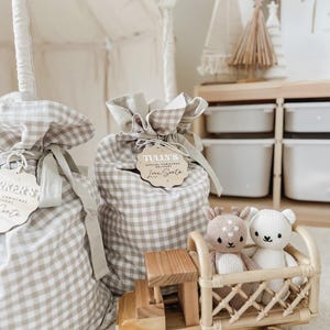 May include: Two gift bags with a tan and white gingham pattern, tied with a ribbon and wooden tag. A wooden toy truck carries a deer and bear plush. A white tent is in the background.