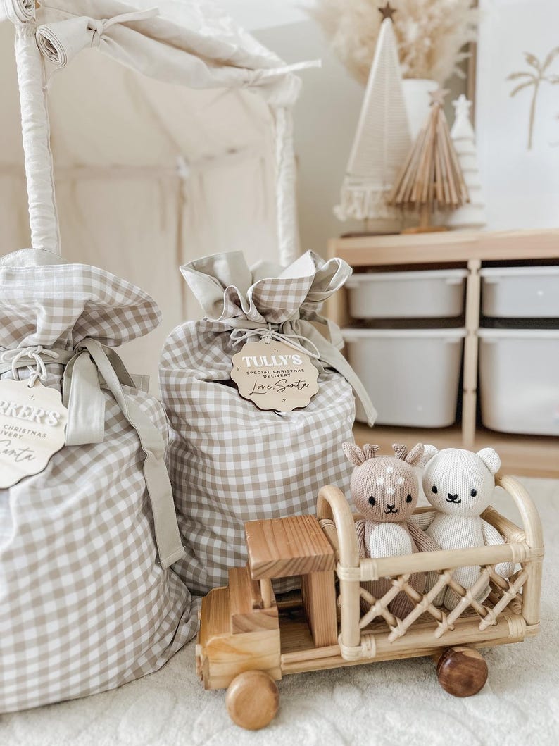 May include: Two gift bags with a tan and white gingham pattern, tied with a ribbon and wooden tag. A wooden toy truck carries a deer and bear plush. A white tent is in the background.