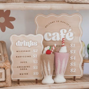 May include: Wooden cafe sign with the text "Welcome to Oaklee's Cafe" and a drinks menu. Two felt milkshakes, one brown and one pink, are displayed in front of the sign. The sign also has the text "EST. 2024".