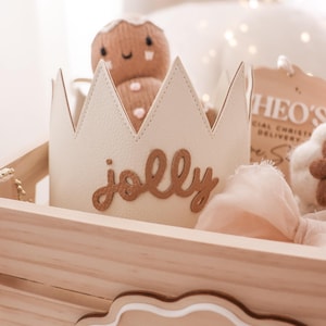 May include: A cream-colored crown with the word "jolly" in brown script is inside a wooden crate. A gingerbread man plush toy and a tag are also in the crate. The crate has a "Merry Christmas" sign.