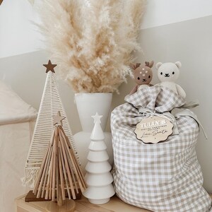 May include: A decorative Christmas display featuring a beige and white gingham gift bag with a wooden tag that reads "Tully's Special Christmas Delivery Love Santa". The display includes a white ceramic tree, a wooden tree, and two stuffed animal toys.
