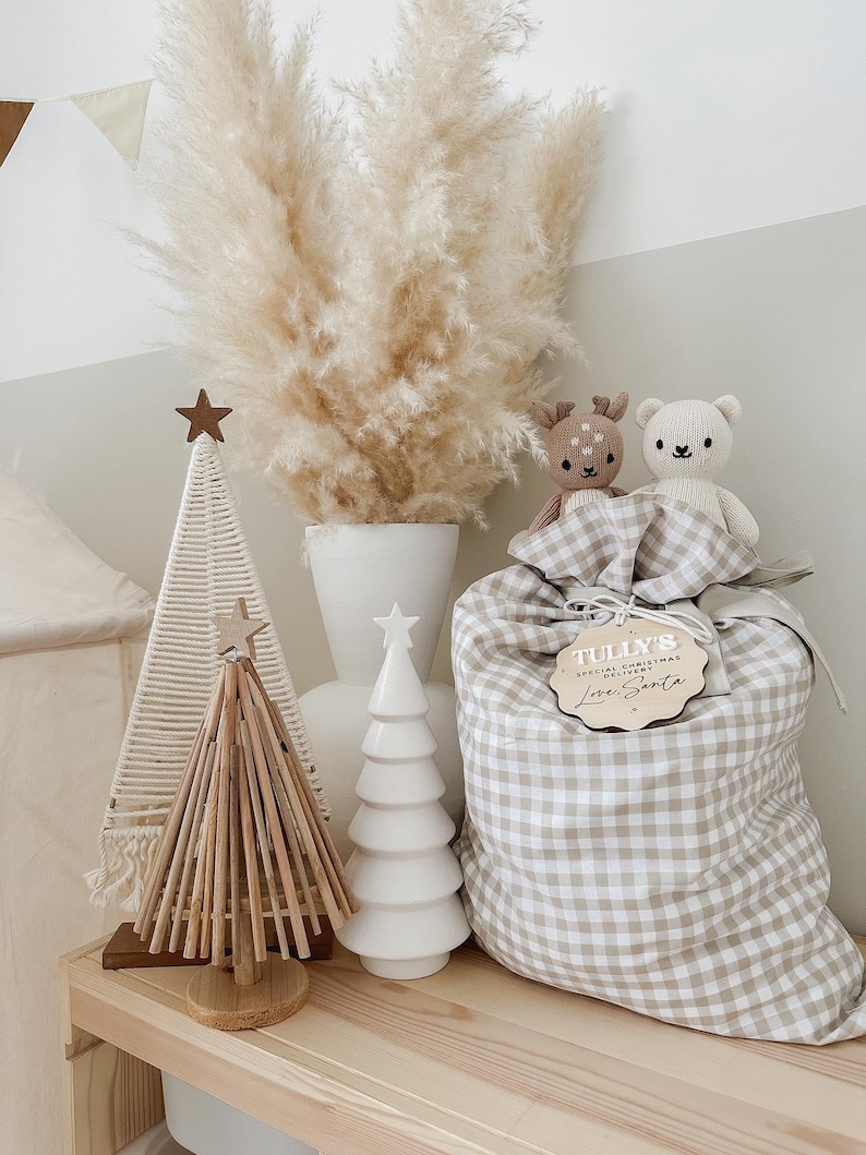 May include: A decorative Christmas display featuring a beige and white gingham gift bag with a wooden tag that reads "Tully's Special Christmas Delivery Love Santa". The display includes a white ceramic tree, a wooden tree, and two stuffed animal toys.