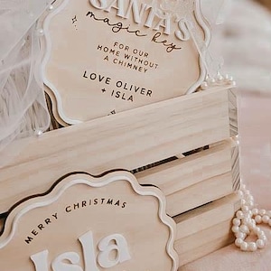 May include: Wooden Christmas ornaments with white lettering and scalloped edges are displayed in a wooden crate. One ornament reads "SANTA'S magic key" and the other says "MERRY CHRISTMAS Isla". A white ribbon and pearl necklace are also visible.