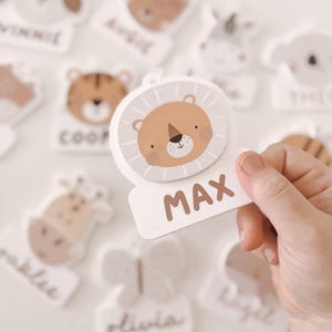 以下が含まれることがあります： 茶色の文字で「MAX」と書かれたライオン型のネームタグが手に持たれています。犬、トラ、キリン、ゾウなど、他の動物をテーマにしたネームタグが背景にあります。タグは薄茶色と白です。