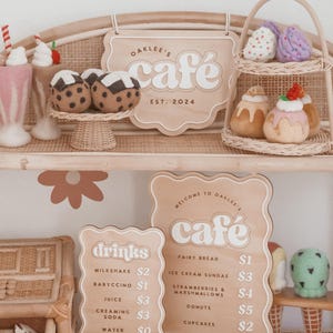 May include: A charming display of felt food and cafe signs. The signs read "Oaklee's Cafe" and list menu items like milkshakes, donuts, and cupcakes. The felt food includes cupcakes, ice cream sundaes, and muffins. The cafe est. 2024.