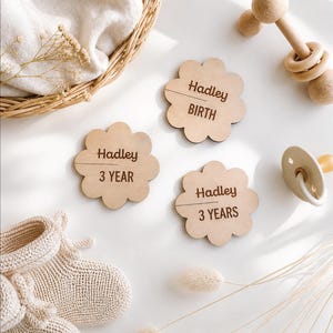 Op de afbeelding: Drie lichtgekleurde houten bloemvormige plaatjes gegraveerd met de naam Hadley en de tekst BIRTH, 3 YEAR en 3 YEARS. De plaatjes liggen naast een paar beige gebreide slofjes, een houten rammelaar en een crèmekleurige fopspeen.