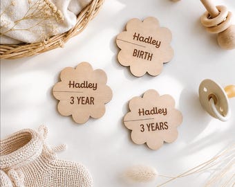 May include: Three light-coloured wooden flower-shaped plaques engraved with the name Hadley and the text BIRTH, 3 YEAR, and 3 YEARS. The plaques are arranged alongside a pair of beige knit booties, a wooden rattle, and a cream-coloured pacifier.
