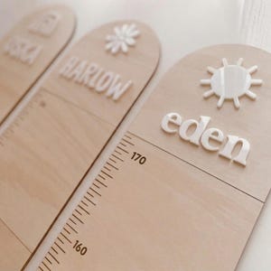 Könnte beinhalten: Drei hölzerne Wachstumstafeln mit Namen und dekorativen Elementen. Jede Tafel hat einen weißen Namen, wie "Eden", und eine Messskala in Zentimetern. Eine Tafel hat eine Sonne, eine andere eine Blume.