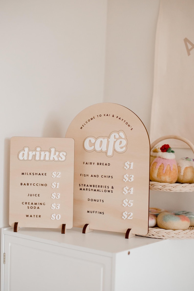 Pu&ograve; includere: Due cartelli in legno con un menu di caffetteria. Il cartello a sinistra dice "drinks" e elenca i prezzi per milkshake, babyccino, succo, soda cremosa e acqua. Il cartello a destra dice "cafe" e elenca i prezzi per pane delle fate, pesce e patatine fritte, fragole e marshmallow, ciambelle e muffin.