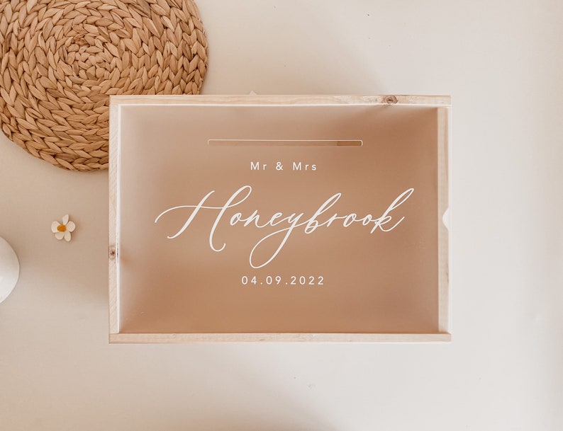 Puede incluir: Una caja de madera con una tapa marr&oacute;n claro y un inserto de acr&iacute;lico transparente. El inserto tiene una ranura para tarjetas y el texto "Mr & Mrs Honeybrook 04.09.2022" en blanco.