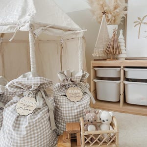 May include: A neutral-toned children's room scene featuring a canvas tent, gift bags, and decorative items. The gift bags are made of a beige and white checkered fabric and have wooden tags. A wooden toy truck carries two stuffed animals. The room is decorated in a minimalist style.