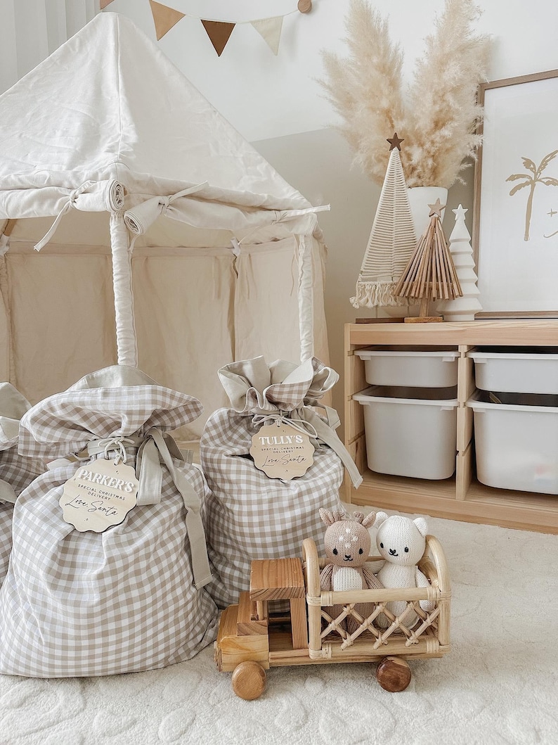 May include: A neutral-toned children's room scene featuring a canvas tent, gift bags, and decorative items. The gift bags are made of a beige and white checkered fabric and have wooden tags. A wooden toy truck carries two stuffed animals. The room is decorated in a minimalist style.