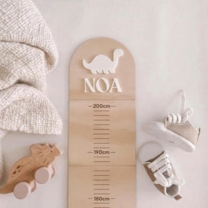 Könnte beinhalten: Eine hölzerne Wachstumstabelle mit einer Dinosaurier-Silhouette und dem Namen "NOA" in Weiß. Die Tabelle misst bis zu 200 cm. Ein Holz-Dinosaurier-Spielzeug und ein Paar kleine Sneaker sind ebenfalls im Bild. Eine beige Decke ist auf der linken Seite drapiert.