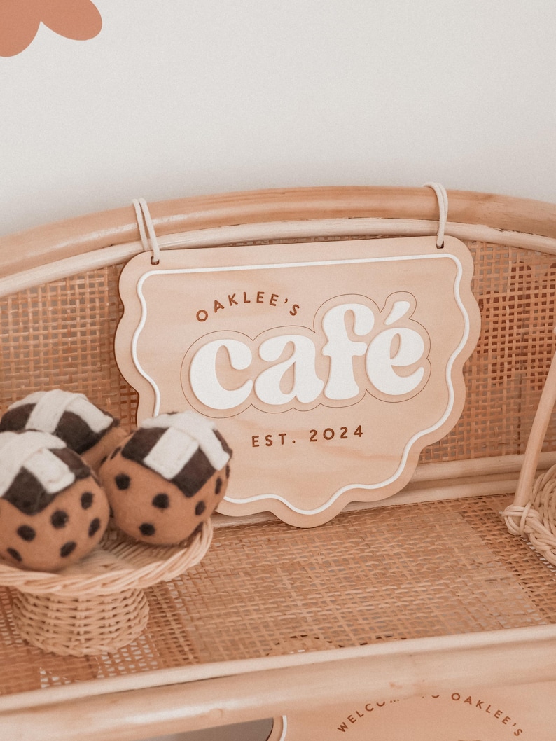 May include: Wooden sign with the text "Oaklee's caf&eacute; EST. 2024" in a playful font. The sign is light brown with a white border and text.  Felt cupcakes with brown and white checkered tops sit on a small wicker tray.