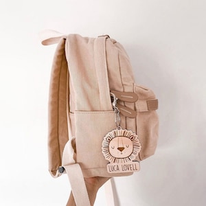 Peut inclure: Un sac à dos en toile beige avec un porte-clés en bois en forme de lion. Le porte-clés porte le nom "Luca Lovell".