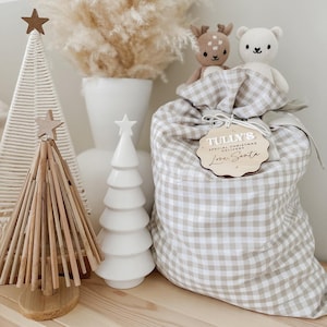May include: A festive Christmas display featuring a beige and white gingham gift bag with a wooden tag that reads "Tully's Special Christmas Delivery Love, Santa." Two small stuffed animals sit atop the bag. Decorative Christmas trees are also present.