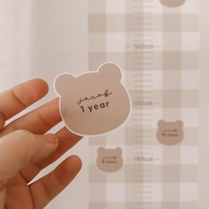 Può includere: Un adesivo beige a forma di orso con il nome "Jacob" e "1 year" in scrittura a mano è tenuto davanti a un'altezza. Il grafico ha un motivo a scacchi beige e bianco con segni di altezza in centimetri.