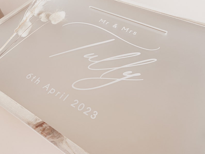 Puede incluir: Una caja de tarjetas de boda de acr&iacute;lico beige claro con un marco de madera blanco. La caja tiene una inscripci&oacute;n blanca que dice "Mr. & Mrs. July 6th April 2023".